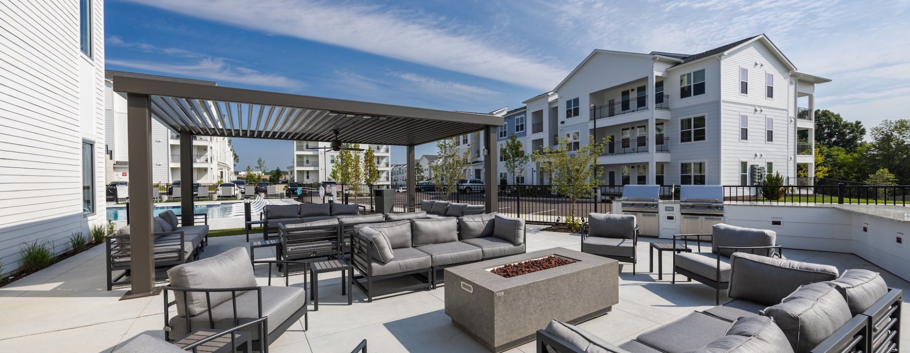 Patio with modern gray seating, fire pit, pergola, and apartment buildings in the background.