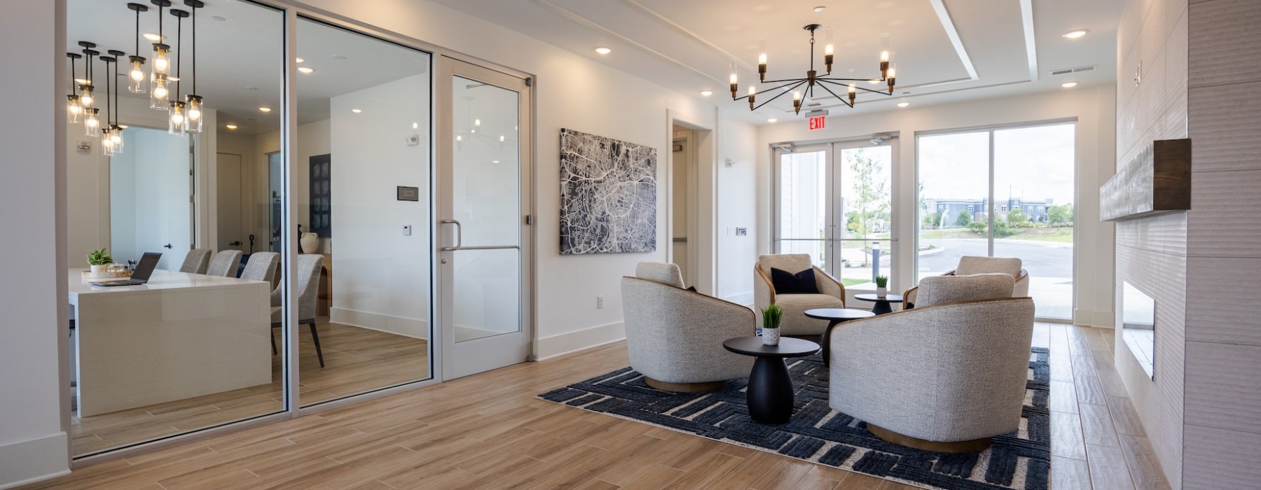 Modern leasing office lounge with round chairs, coffee tables, and chandelier lighting next to a glass-walled meeting room.